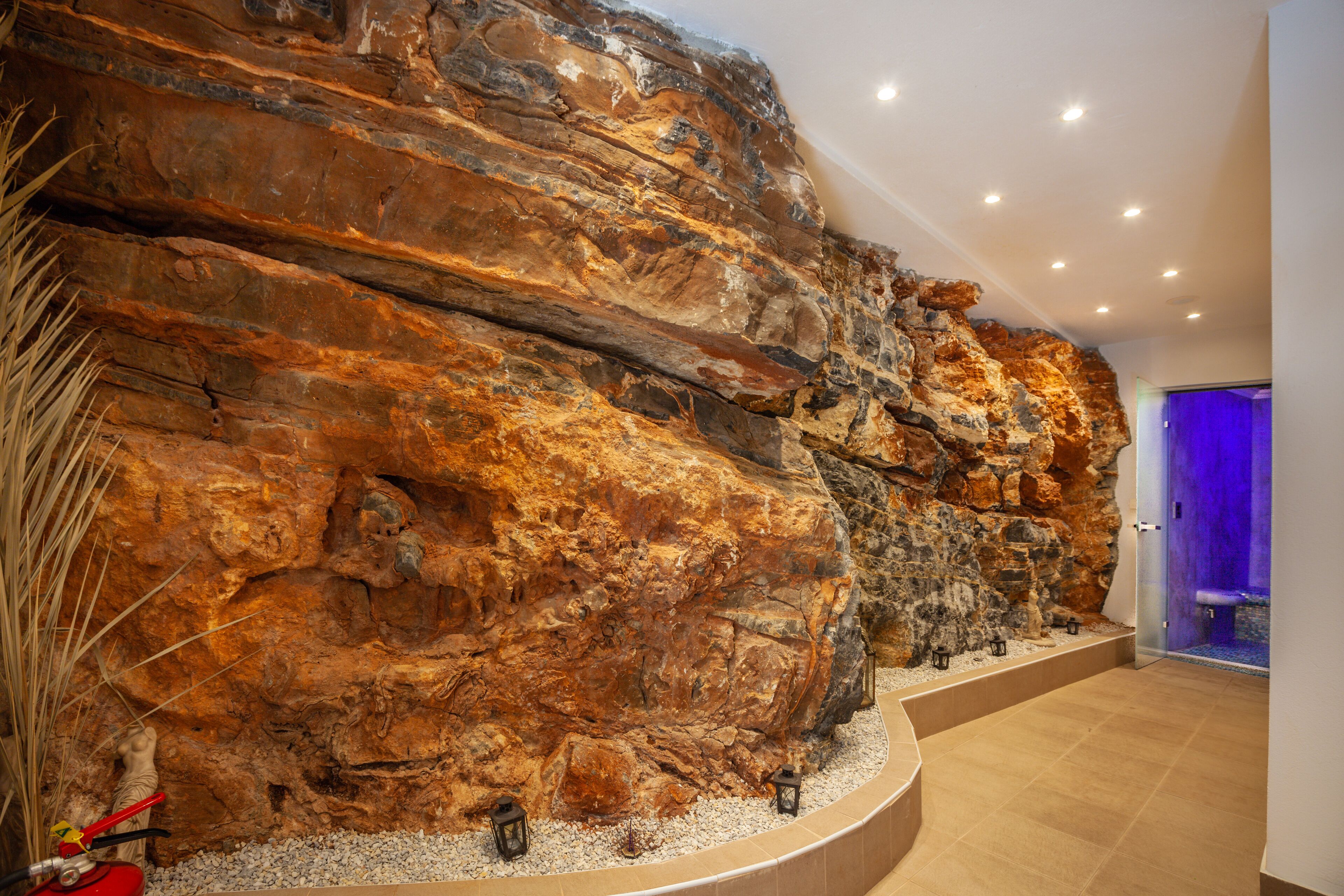 Ein modernes Interieur mit einer großen Steinwand, einem gewundenen Pfad und einer Tür zu einem Raum mit violetter Beleuchtung.