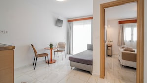 A modern hotel room with a bed, a small table with chairs, a TV mounted on the wall, and a bathroom visible through an open door.