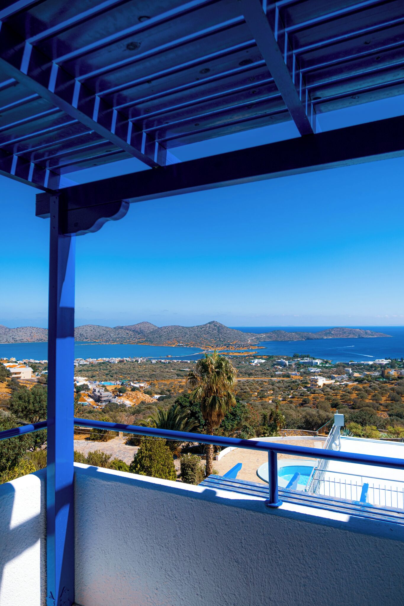 A balcony with a view of a coastal landscape, including a swimming pool and distant hills.