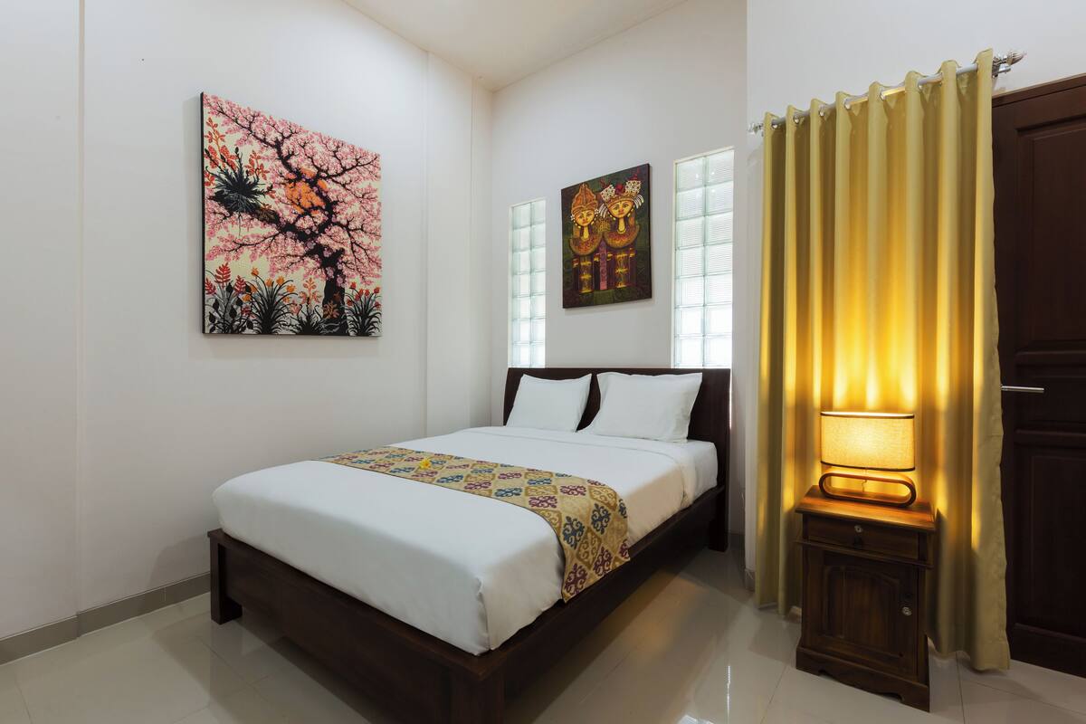 Una cama bien tendida con una colcha estampada, dos cuadros colgados en la pared, una lámpara de noche y una ventana con cortinas.