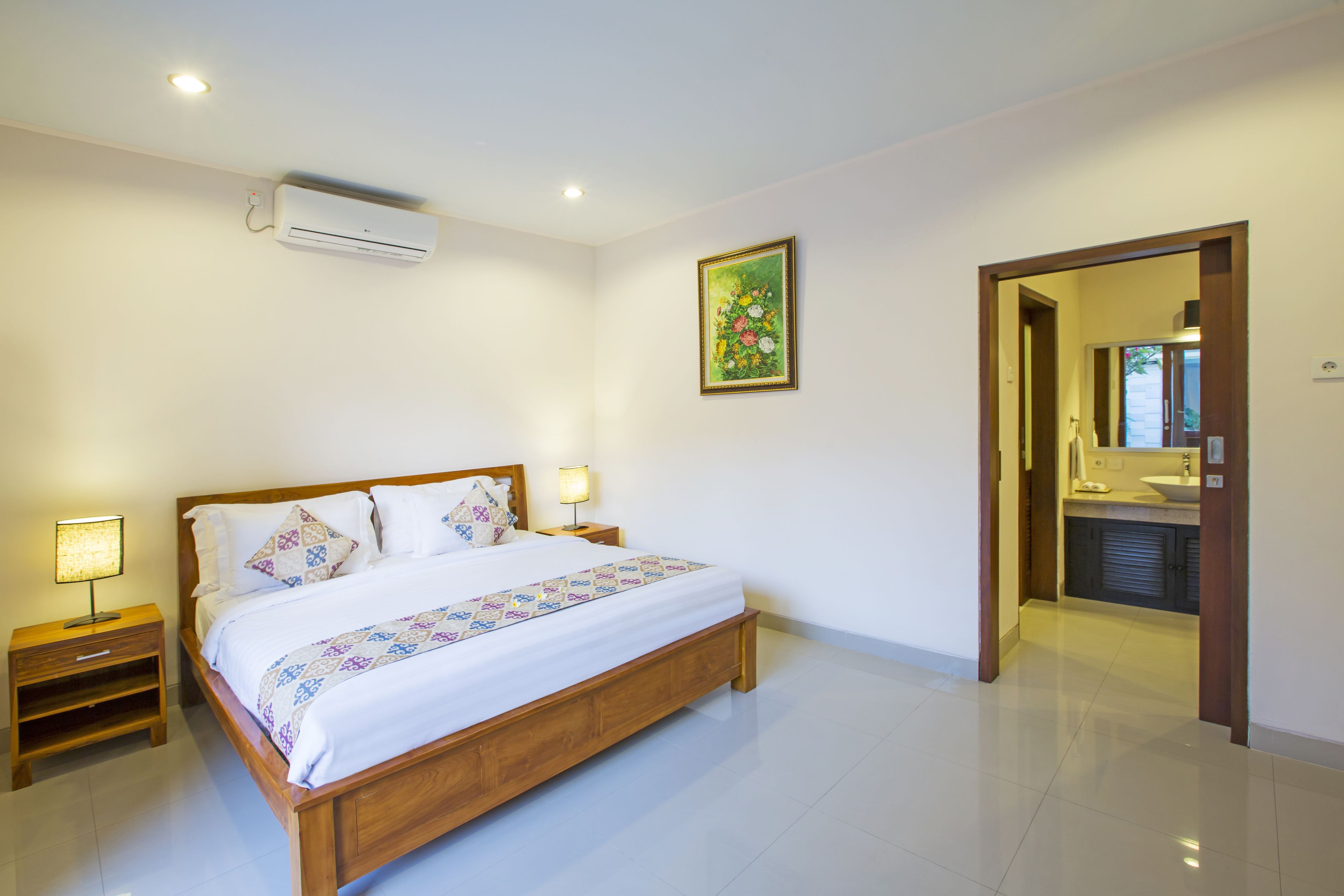 A bedroom with a wooden bed, bedside tables, an air conditioner, a painting, and a bathroom visible through an open door.