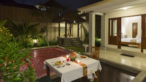 Un coin repas avec une table dressée pour deux, une petite piscine parsemée de pétales de fleurs rouges, et une chambre privée avec un lit et une salle de bain visibles à travers une porte ouverte.