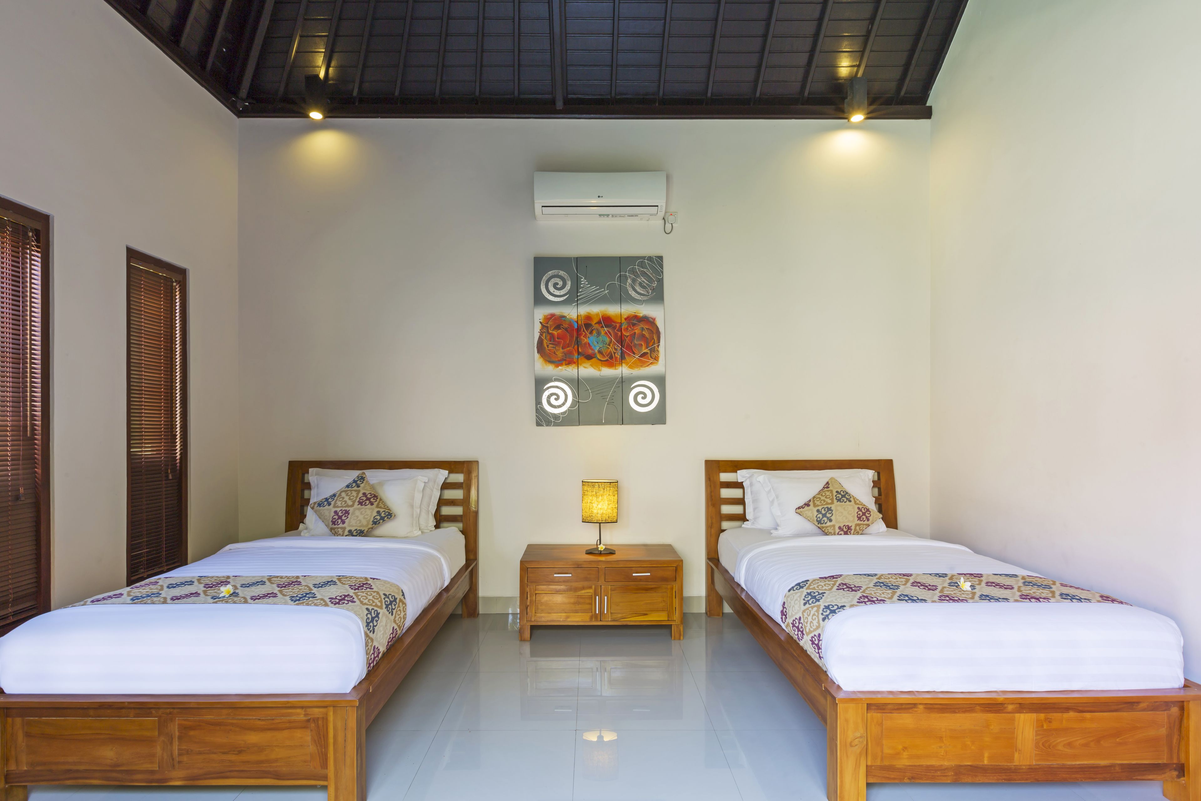 A hotel room with two wooden beds, a nightstand with a lamp, an air conditioning unit, and artwork on the wall.