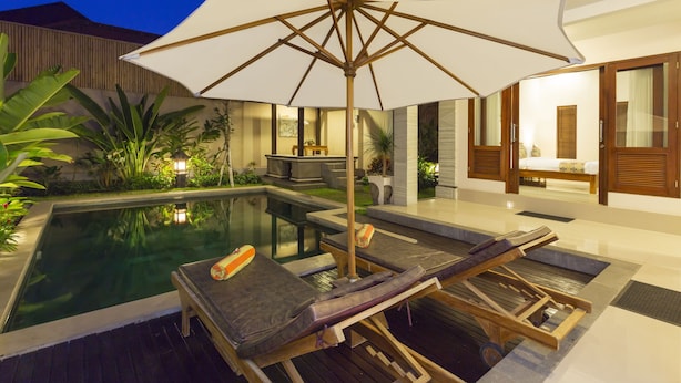 Un espace piscine doté de chaises longues et d’un grand parasol, entouré de plantes et bénéficiant d’un éclairage intérieur agréable.