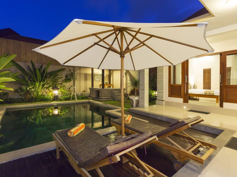 Área de piscina com espreguiçadeiras e um grande guarda-sol, cercada por plantas e um interior bem iluminado.