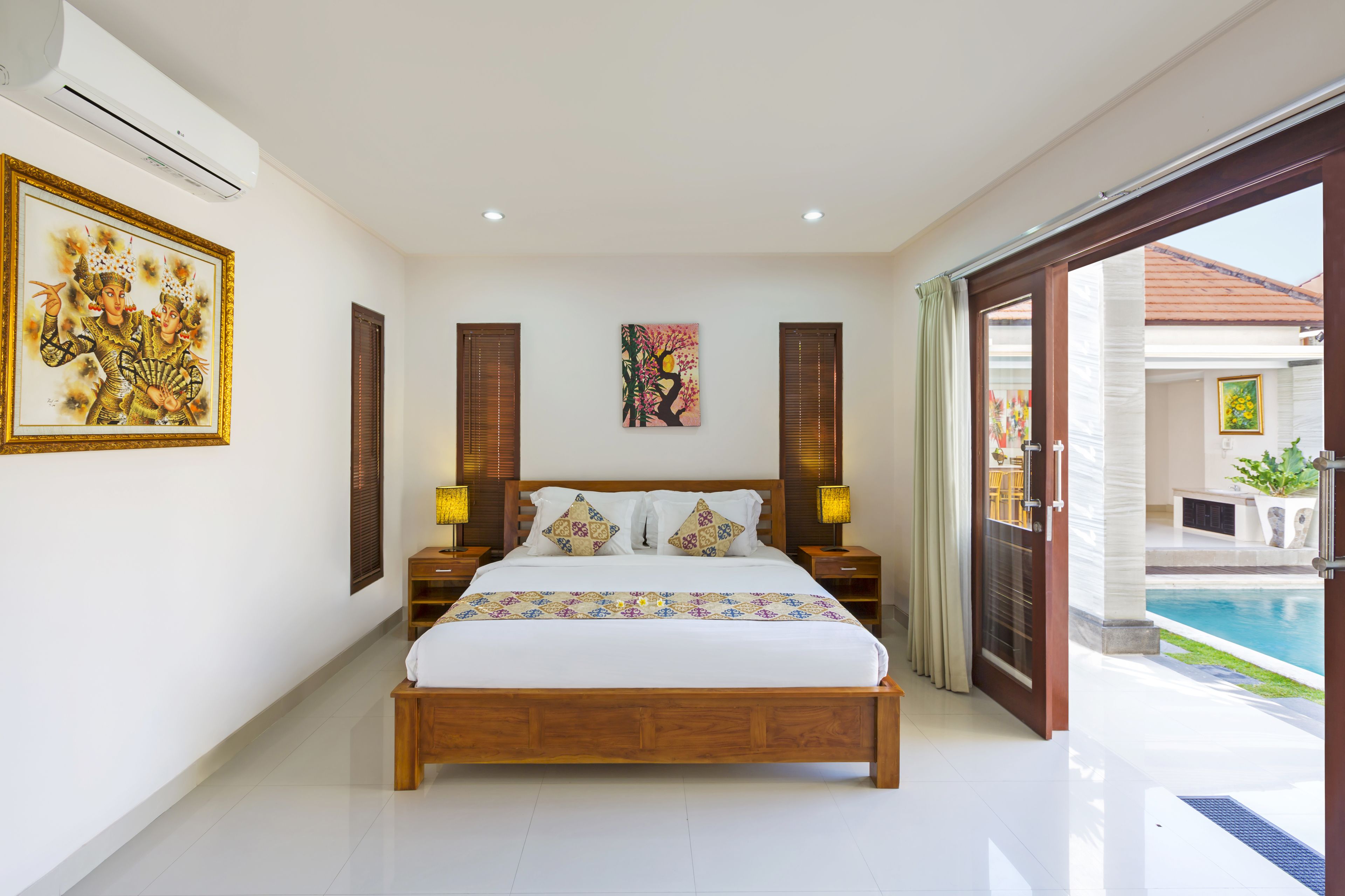 A spacious bedroom with a large bed, two bedside tables with lamps, a wooden headboard, and a view of a swimming pool through an open door.