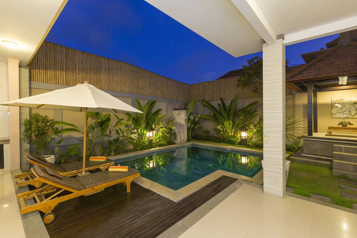 Un área de piscina moderna al aire libre con sillones de descanso y una sombrilla, rodeada de exuberante vegetación y una terraza de madera.