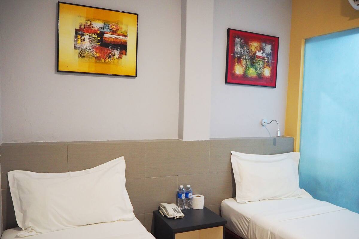 Een hotelkamer met twee bedden, een nachtkastje met telefoon en flessen water, en twee abstracte schilderijen aan de muur.