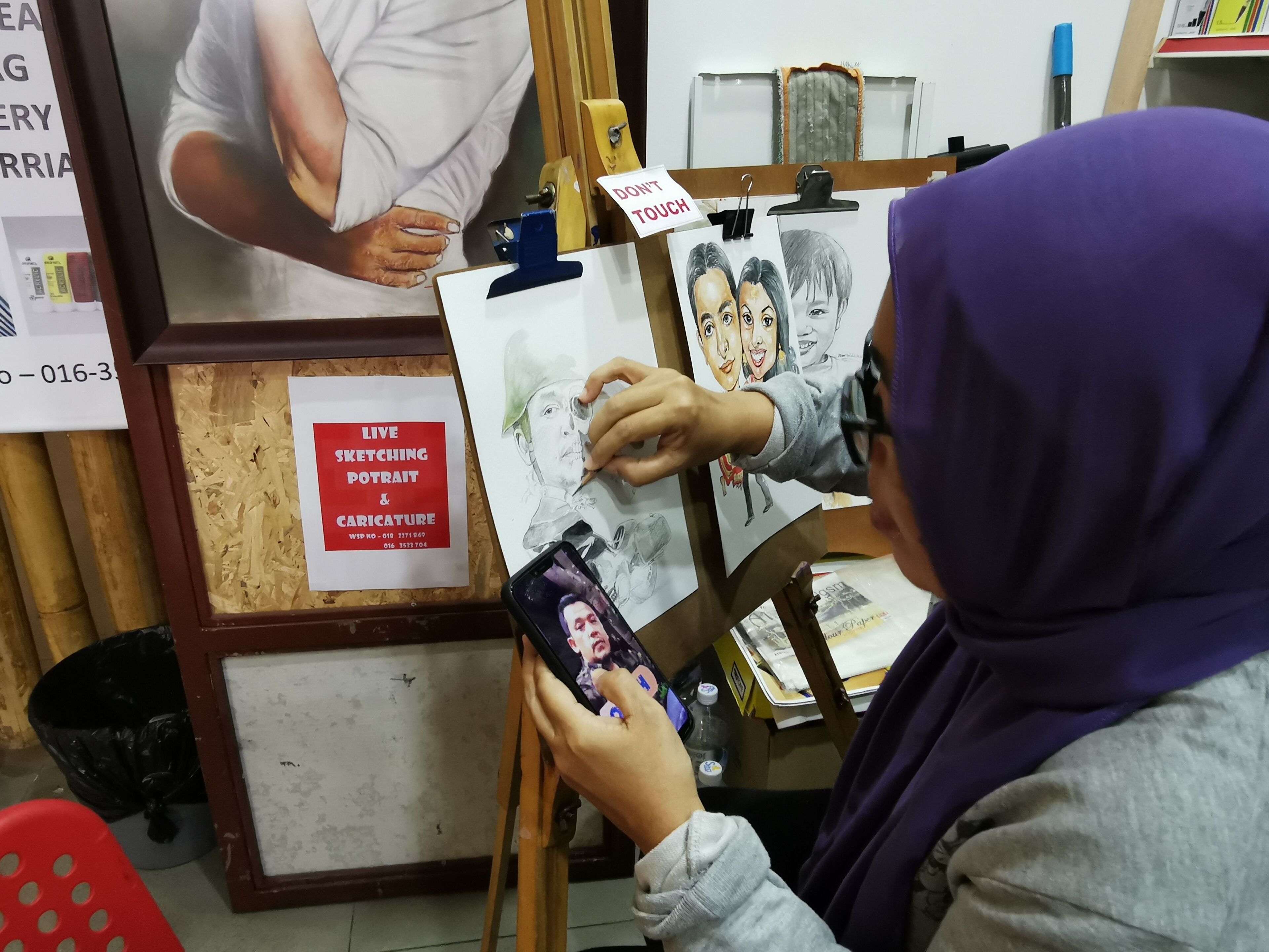 A person sketching on a drawing board with a 'DON'T TOUCH' sign, surrounded by art supplies and a framed poster advertising live sketching and caricature services.