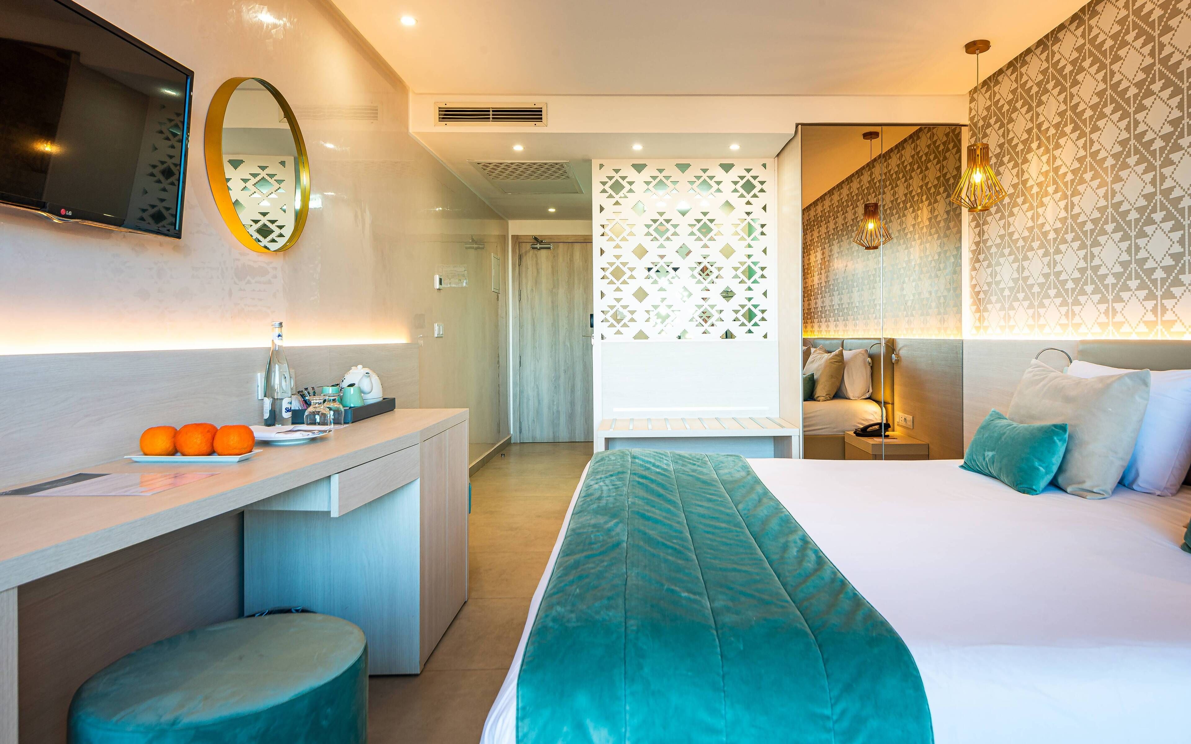 A modern hotel room with a bed, a desk, a mirror, and a bathroom visible through a partition.