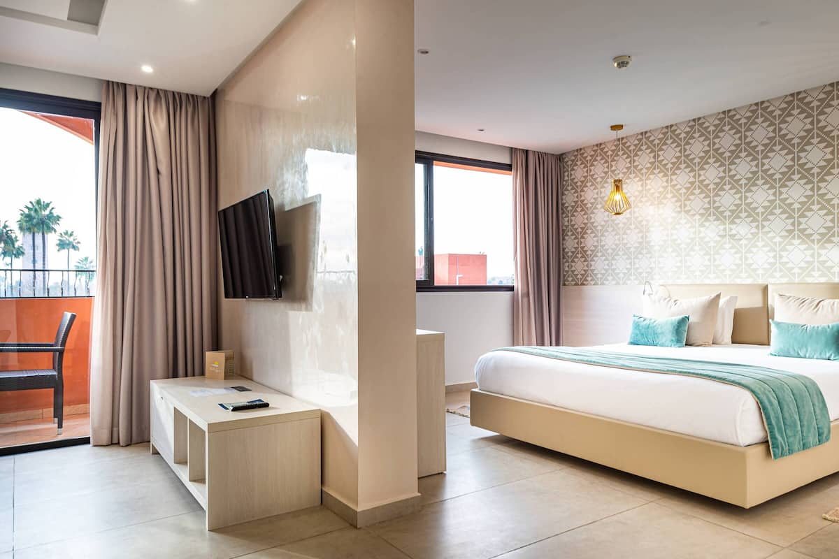 Habitación de hotel con cama, televisor montado en la pared, escritorio y balcón con una silla.
