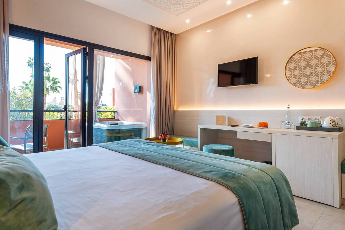 Habitación de hotel con cama, escritorio, televisión y balcón con vista a palmeras.