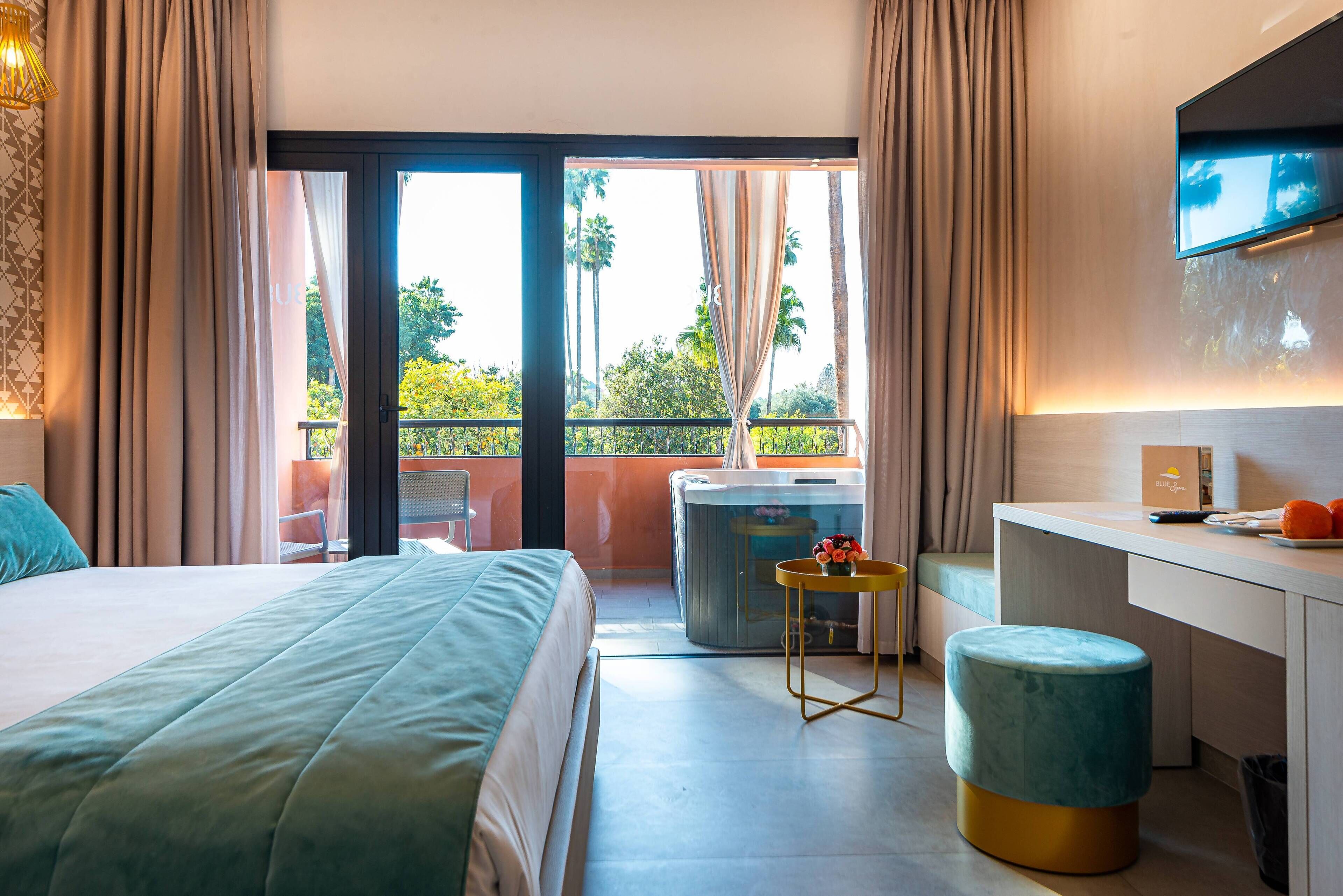 A hotel room with a bed, a desk, a chair, a small table with flowers, a trash bin, and a view of the outdoors.