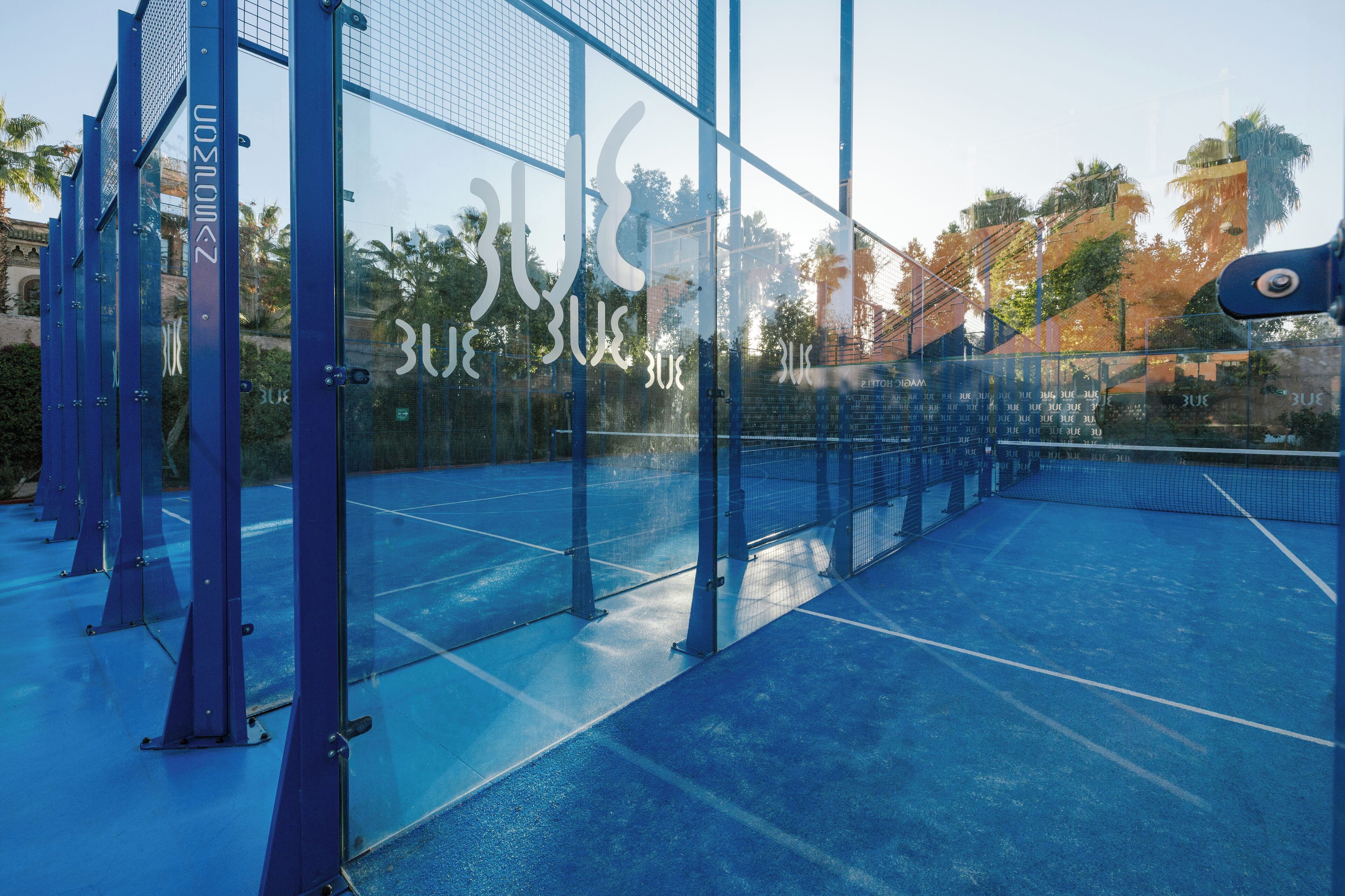 Una cancha de tenis con paredes de vidrio y piso azul, rodeada de estructuras metálicas azules.