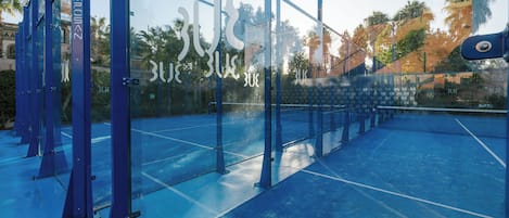 Una cancha de tenis con paredes de vidrio y piso azul, rodeada de estructuras metálicas azules.