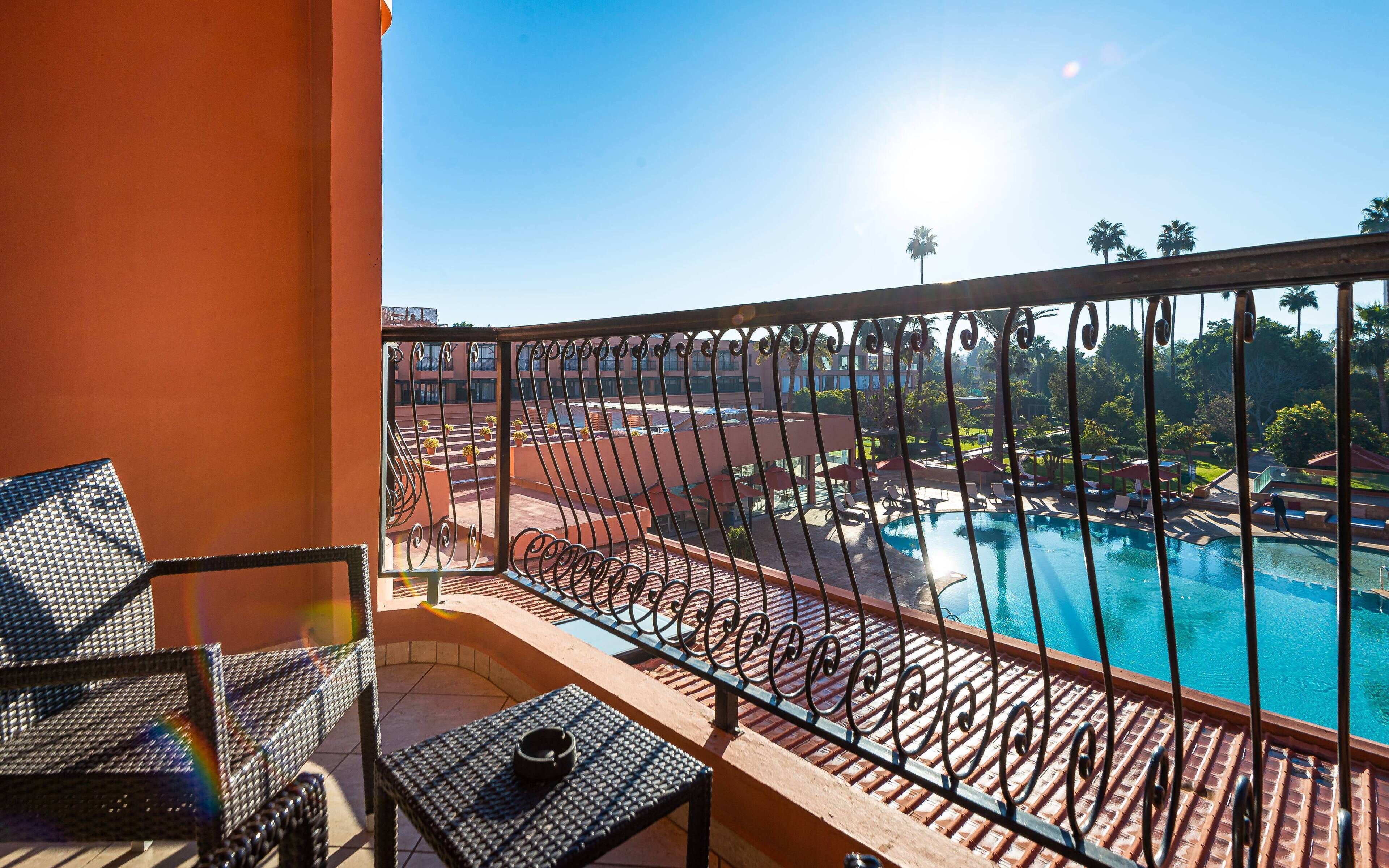 A balcony with a view of a swimming pool, lounge chairs, and a sunlit outdoor area.