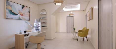 A modern salon with a central reception desk, two chairs, a large abstract painting, and a skylight.