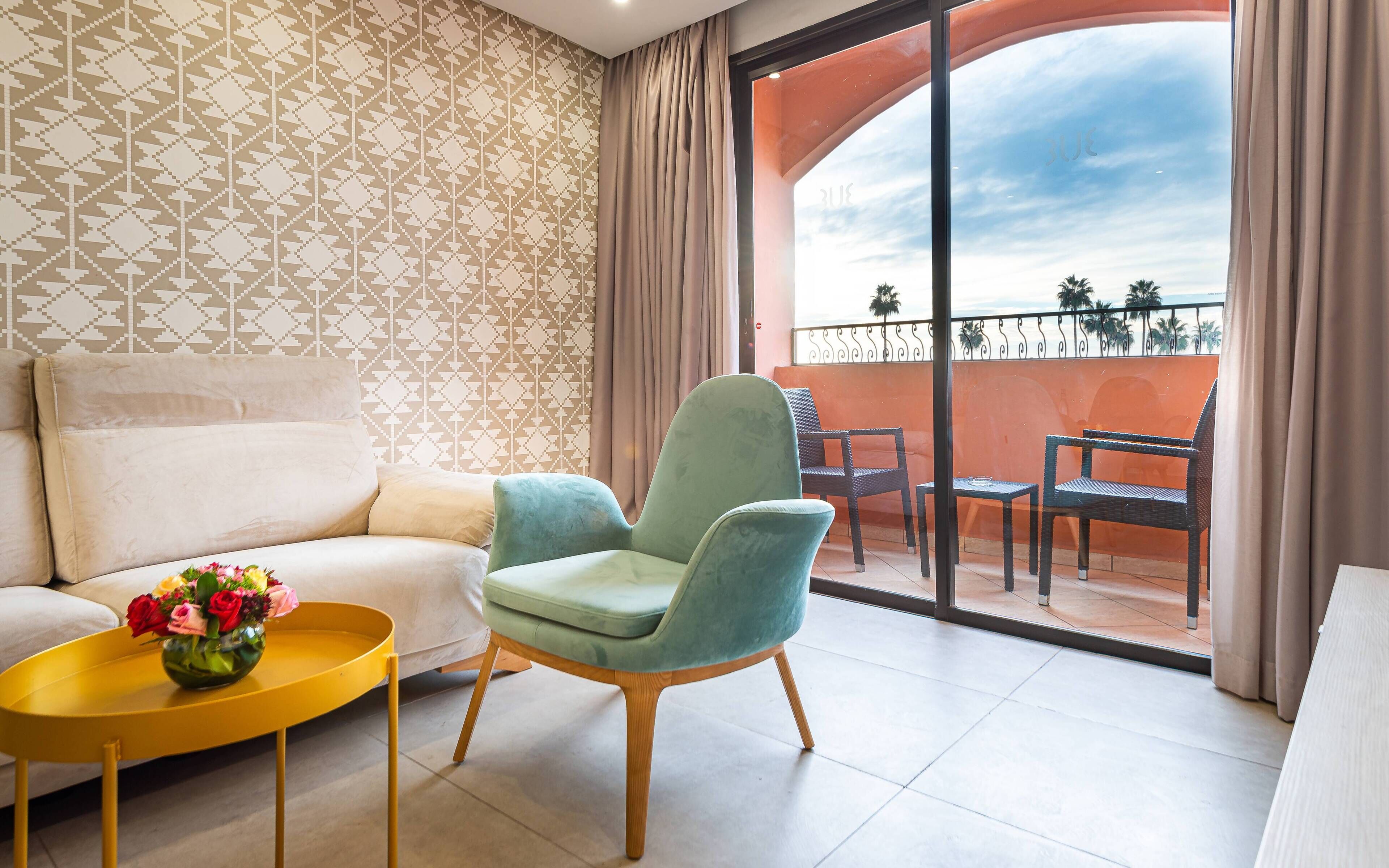 A modern living room with a sofa, a green armchair, a yellow coffee table with a vase of flowers, and a balcony with outdoor furniture.