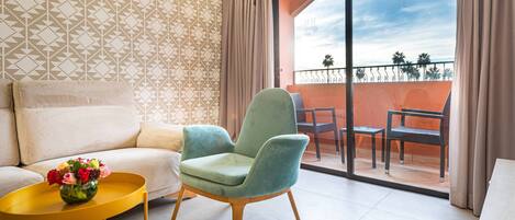 A modern living room with a sofa, a green armchair, a yellow coffee table with a vase of flowers, and a balcony with outdoor furniture.
