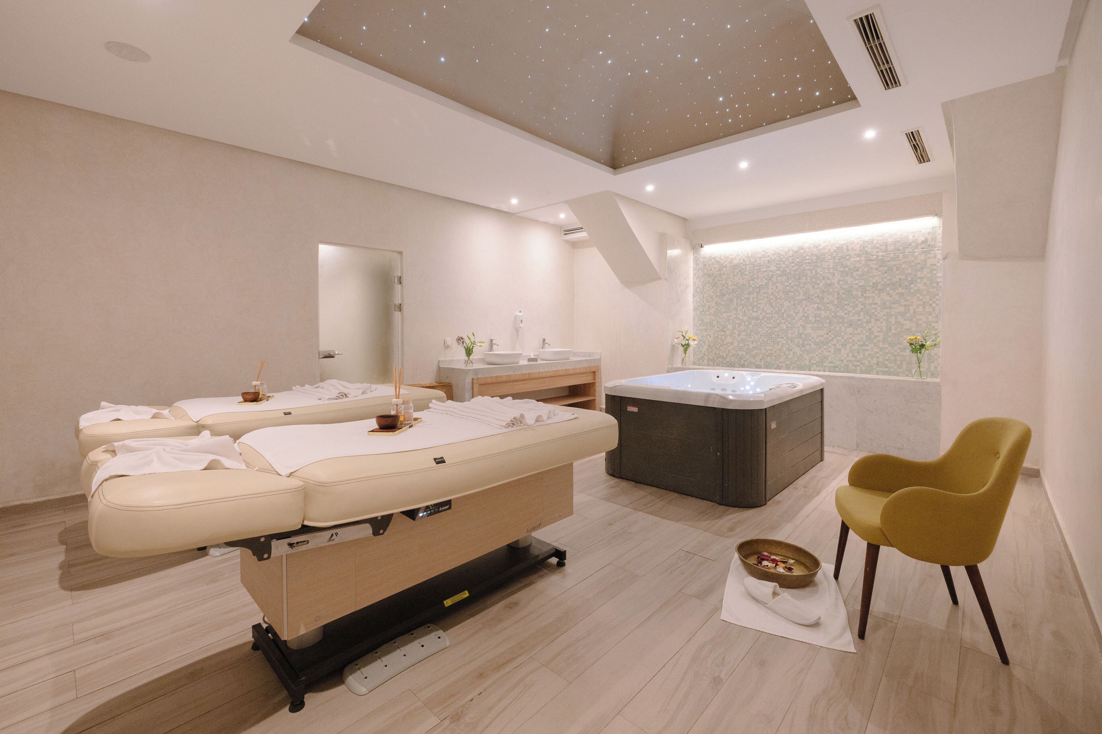 A modern spa room with a massage table, a hot tub, and a yellow chair.