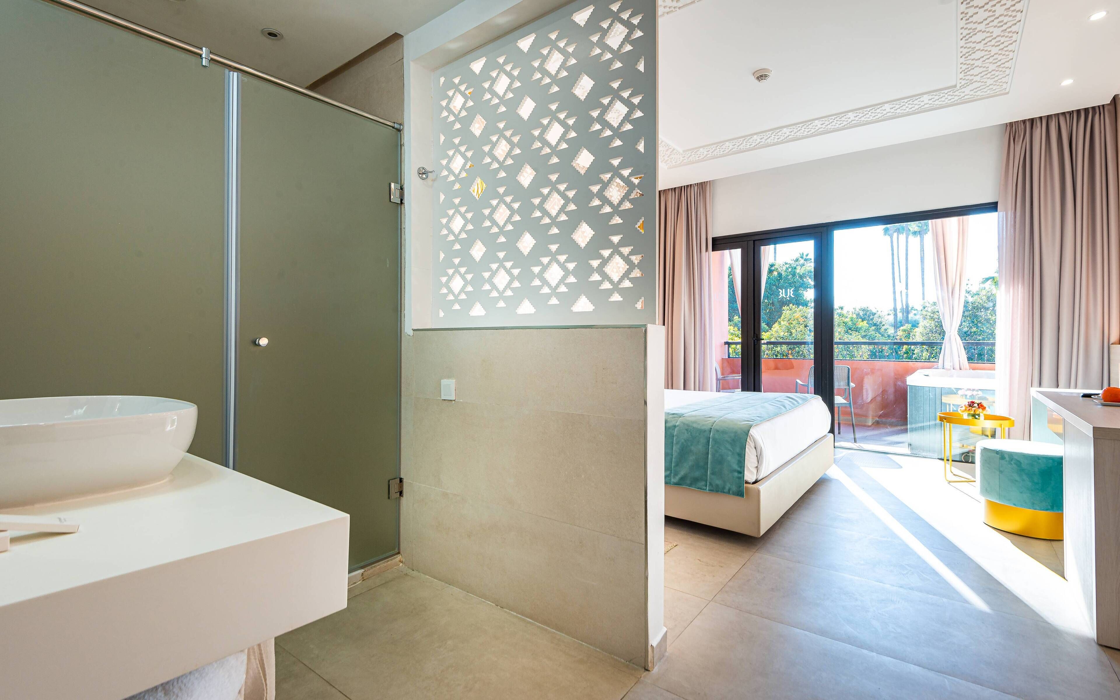 A modern hotel room with a glass shower enclosure, a large bed, a balcony with a view, and a separate bathroom area.