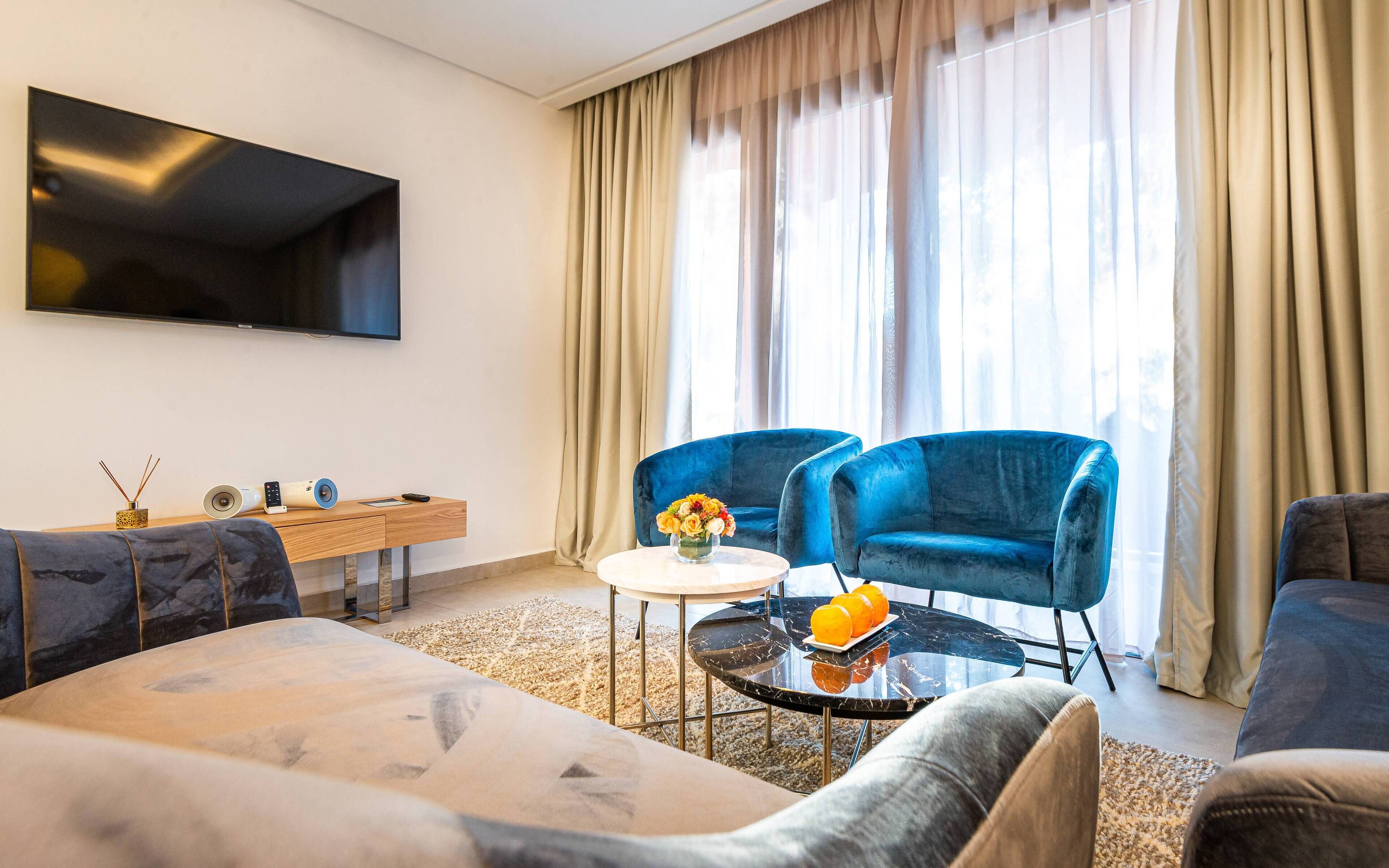 A modern living room with a flat-screen TV, two blue armchairs, a sofa, a coffee table with a vase of flowers, and a wooden side table with a lamp and a speaker.