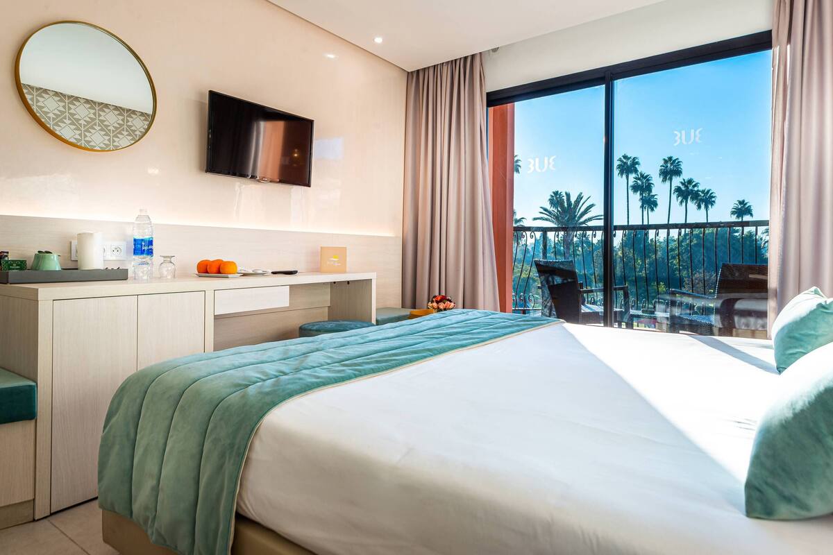 Habitación de hotel con cama, escritorio, espejo y vistas a palmeras a través de amplias ventanas.
