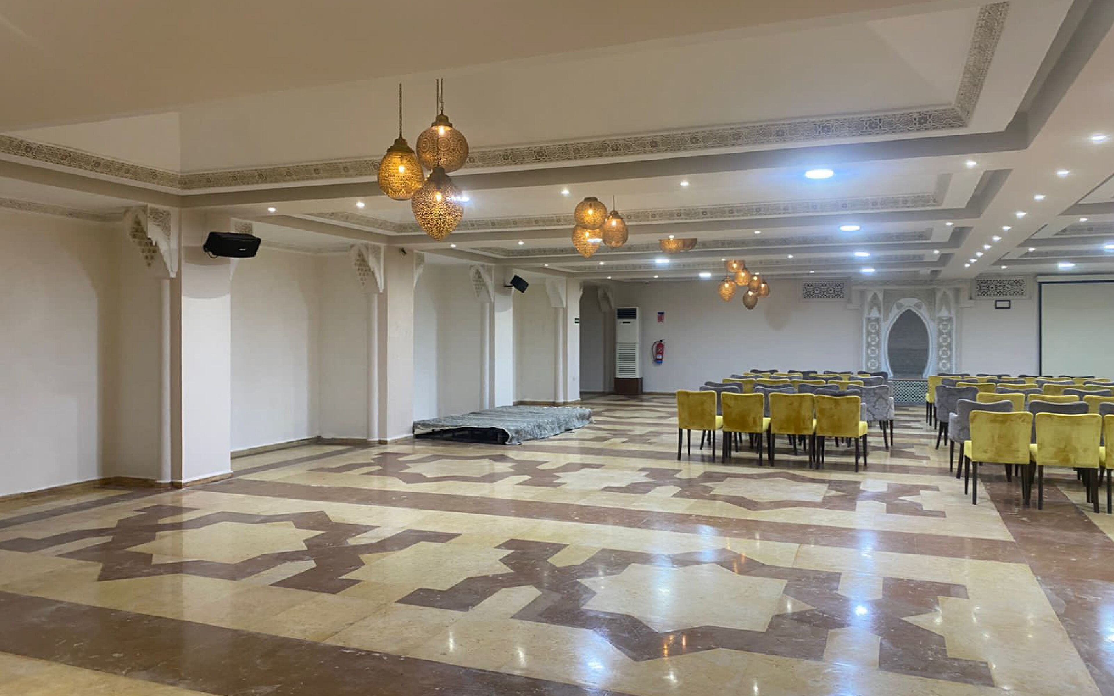 A spacious hall with patterned flooring, rows of yellow chairs, and hanging pendant lights.