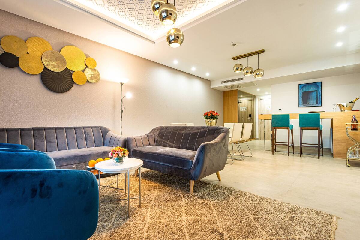 Una sala de estar moderna con un sofá, dos sillones, una mesa de centro y una alfombra. Se observa un comedor con una barra y sillas al fondo.