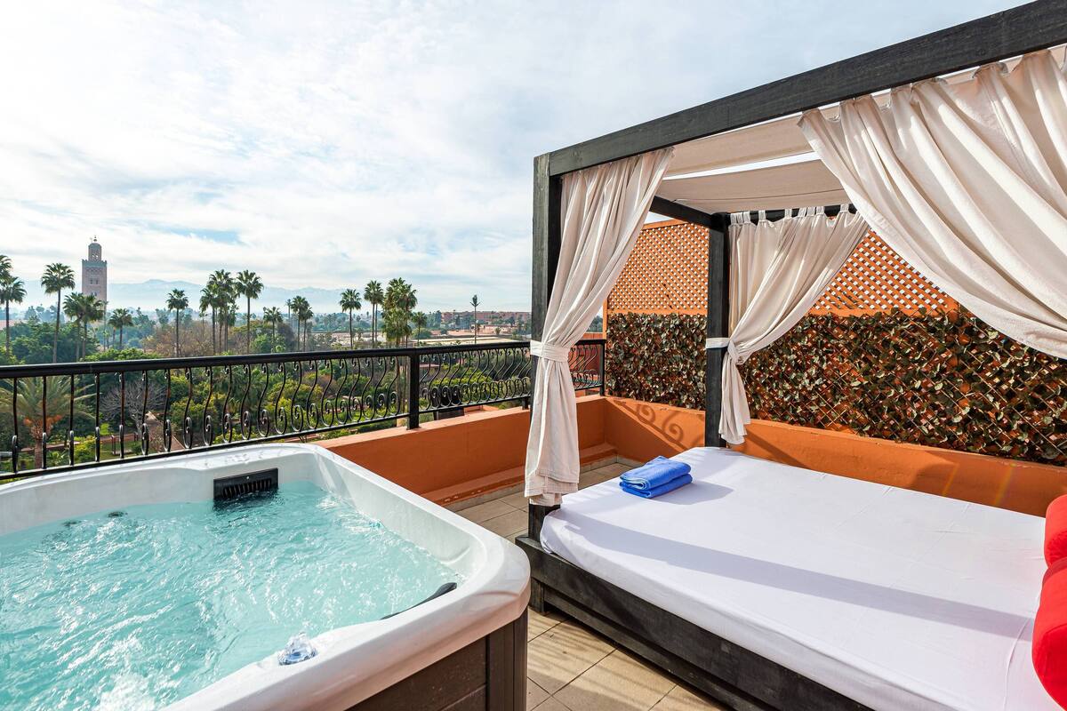 Un balcón con un jacuzzi, una cama y vistas a palmeras y una torre distante.