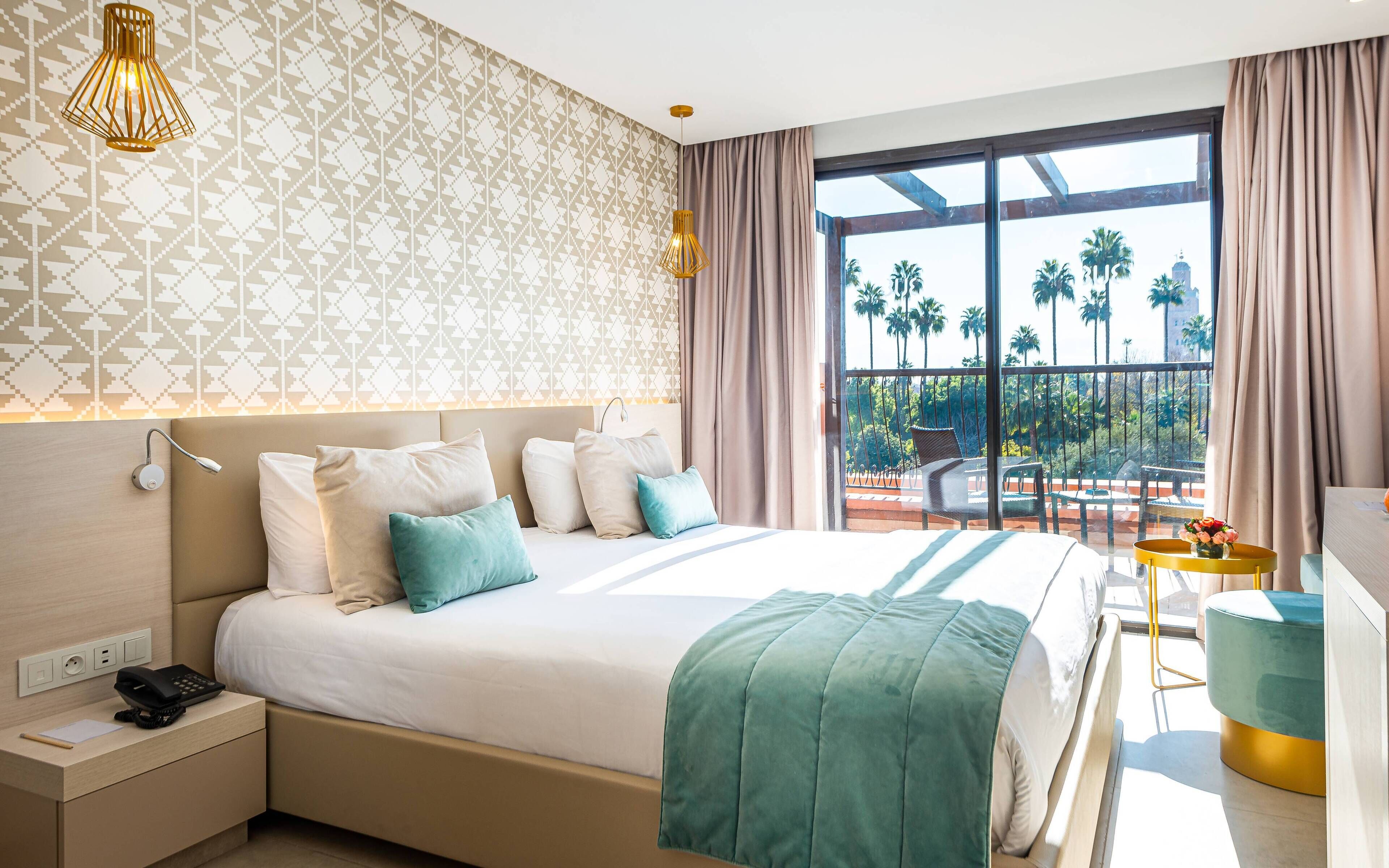 A modern hotel room with a large bed, a bedside table with a phone, and a view of palm trees through large windows.