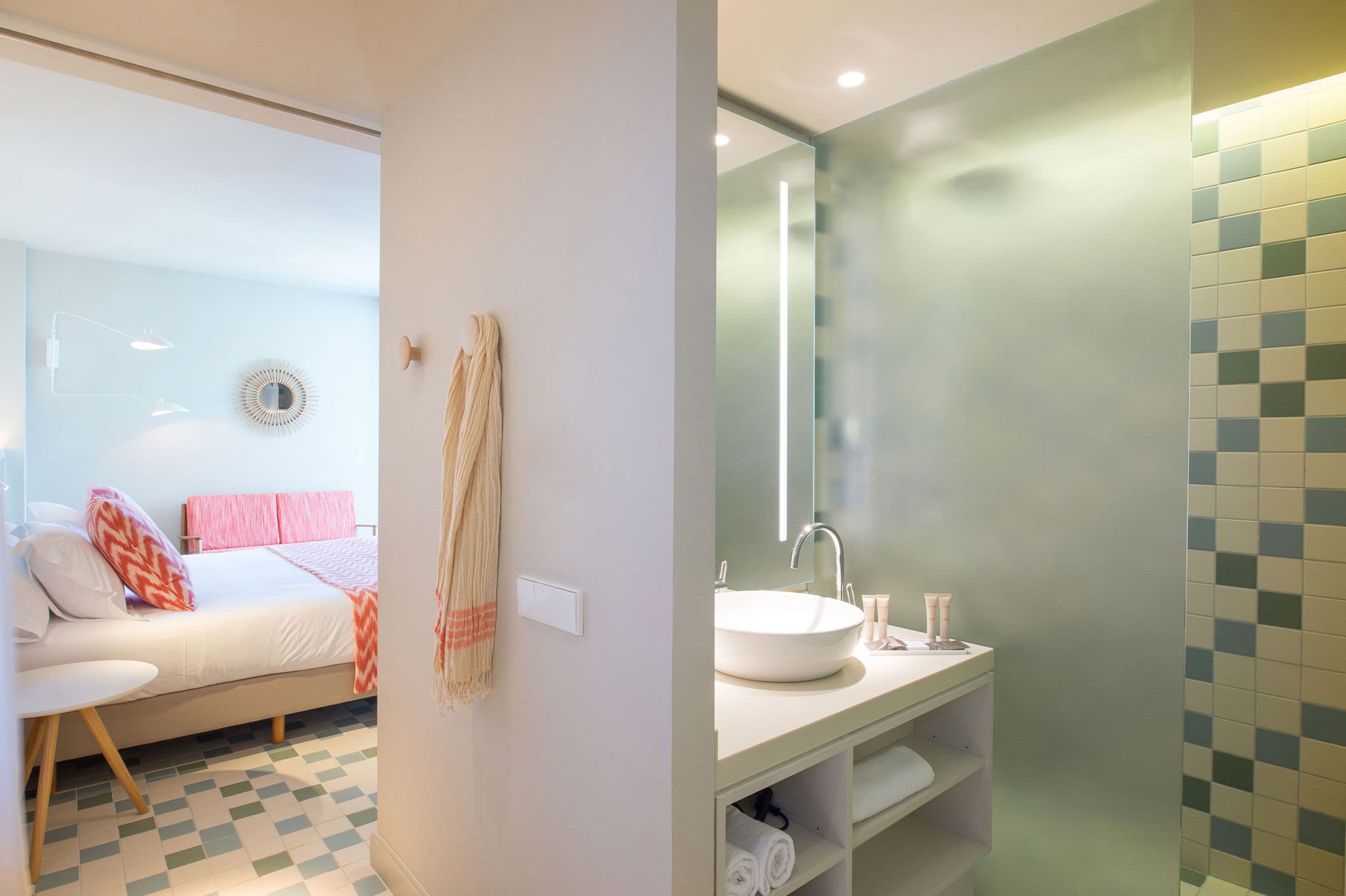 A modern bedroom with a bed, bedside table, and a wall-mounted mirror. The bathroom features a sink, mirror, and tiled walls.