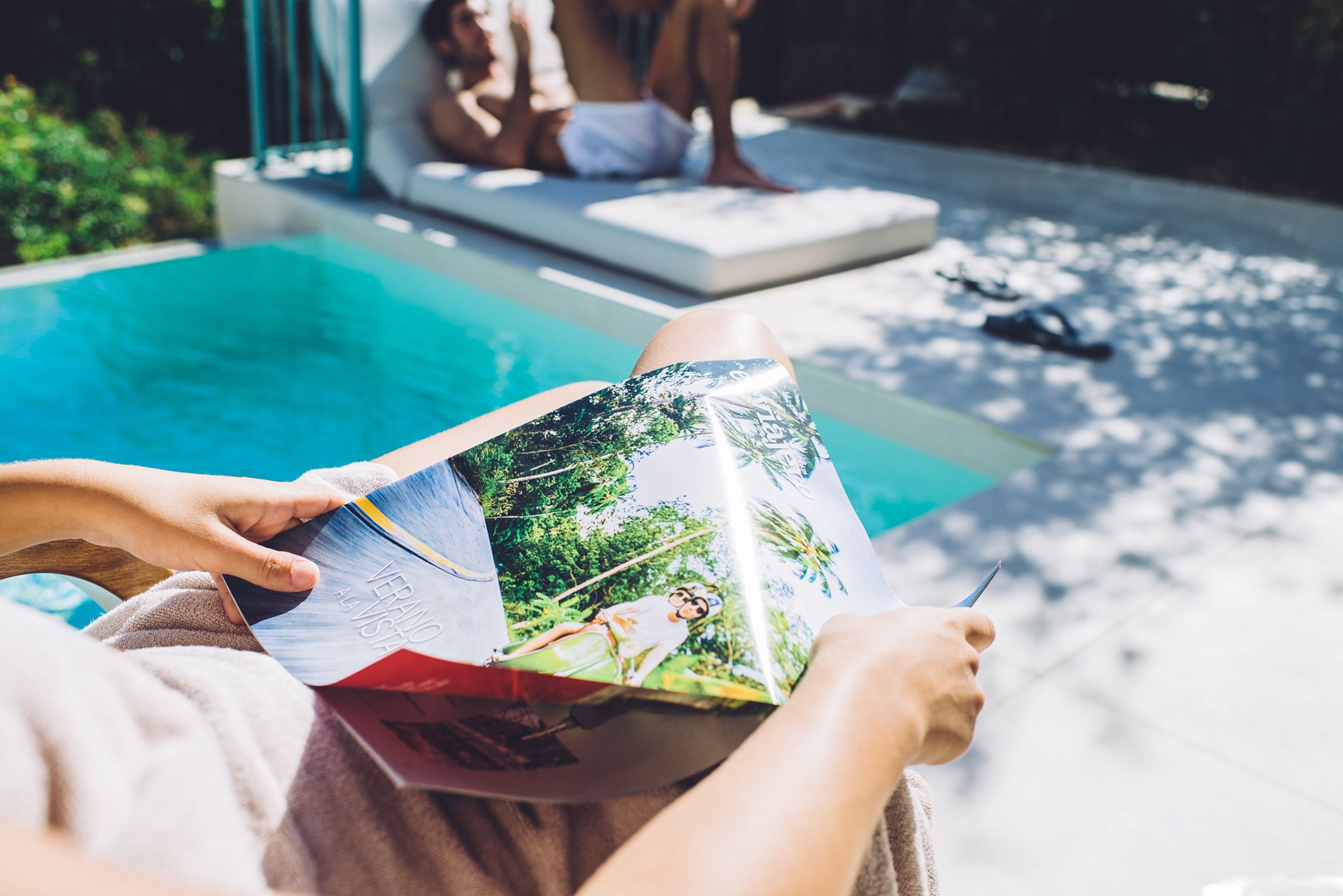 A person reading a magazine by a pool with a clear view of the water and a lounge area in the background.