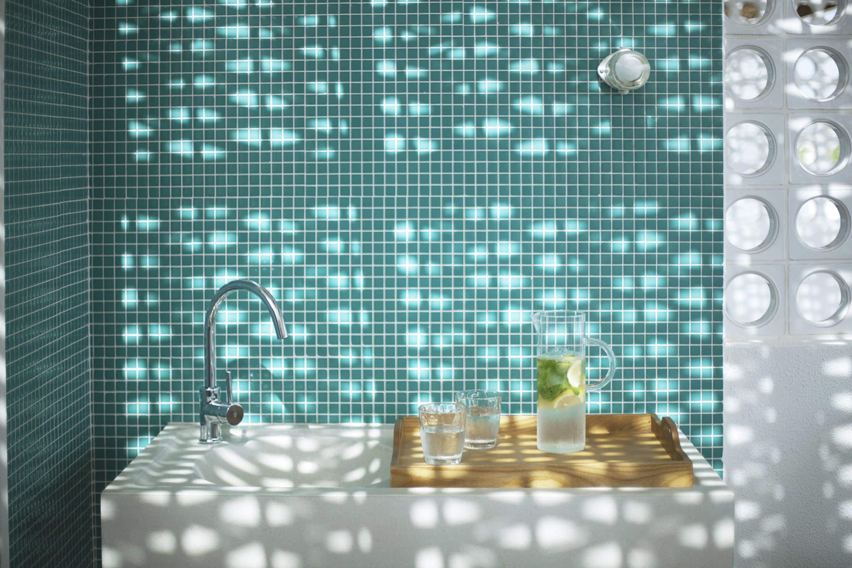 A bathroom with a sink, faucet, and a tiled wall. There are three glasses on a wooden tray.