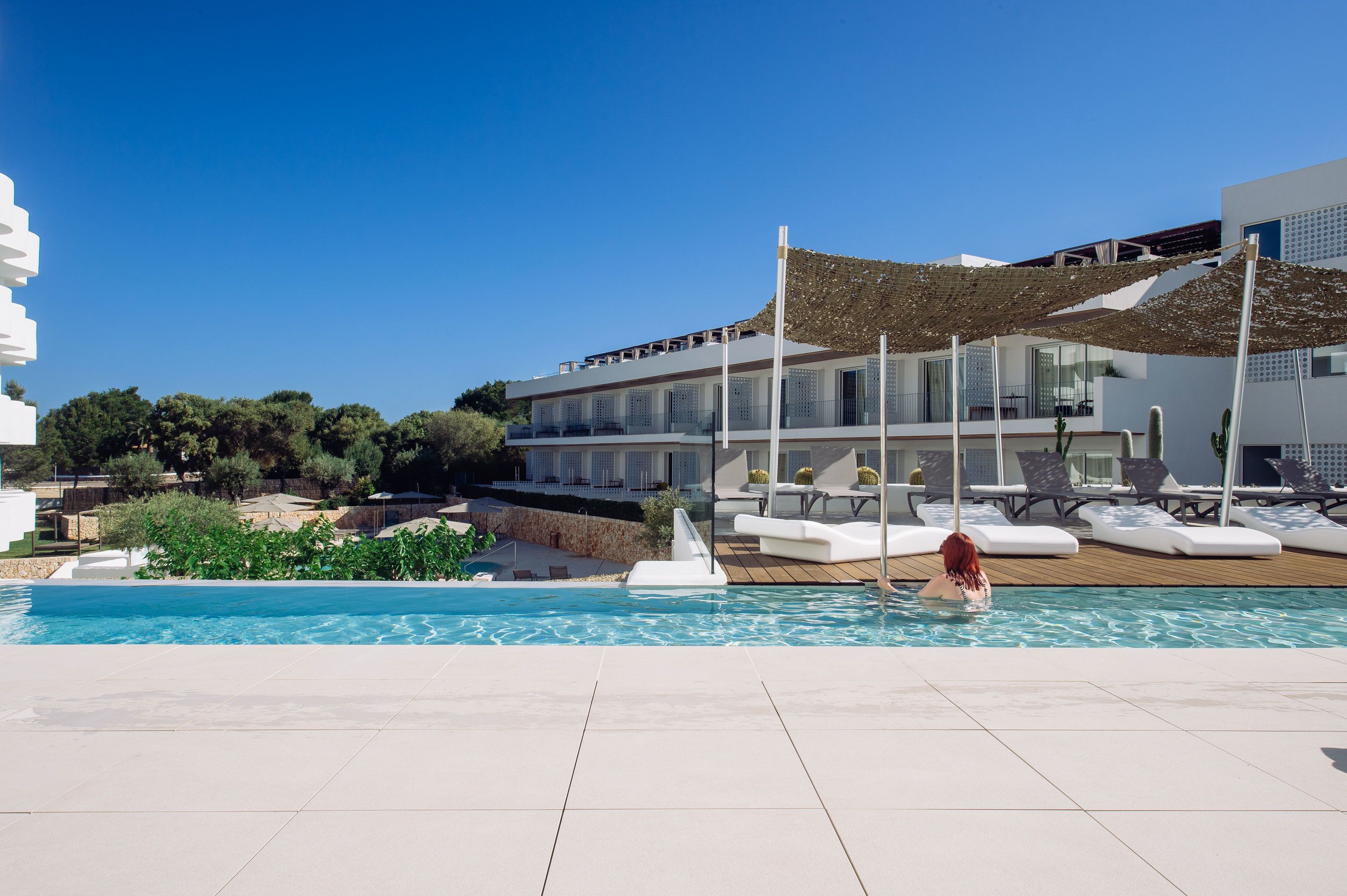 Un área de piscina moderna con un cielo azul despejado, una piscina, sillas de descanso y un edificio con balcones.