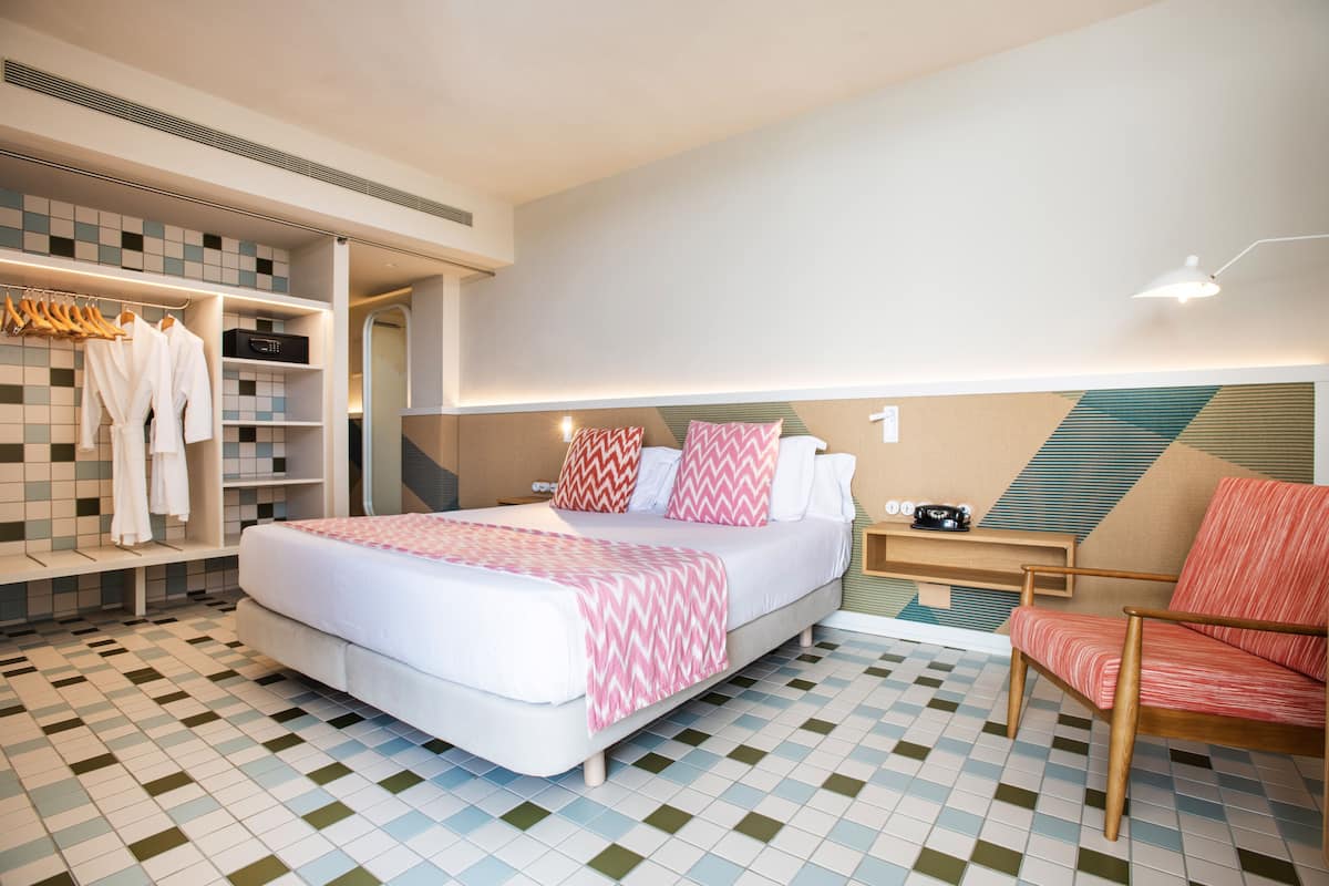 Una habitación de hotel moderna con una cama grande, una zona de estar con una silla, un tocador con espejo y un armario con batas.
