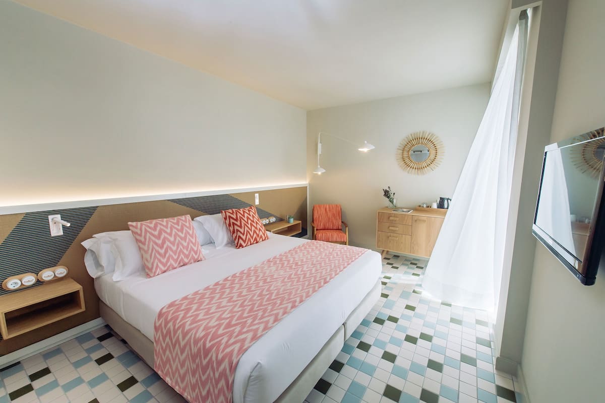 Habitación de hotel con cama, mesita de noche, lámpara y vistas al exterior.