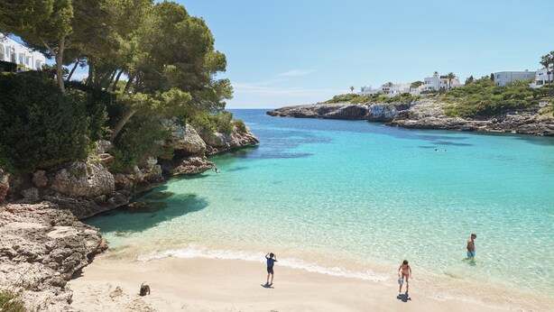Ein Küstenstrand mit klarem türkisblauem Wasser, felsigen Klippen und Gebäuden am Hang.