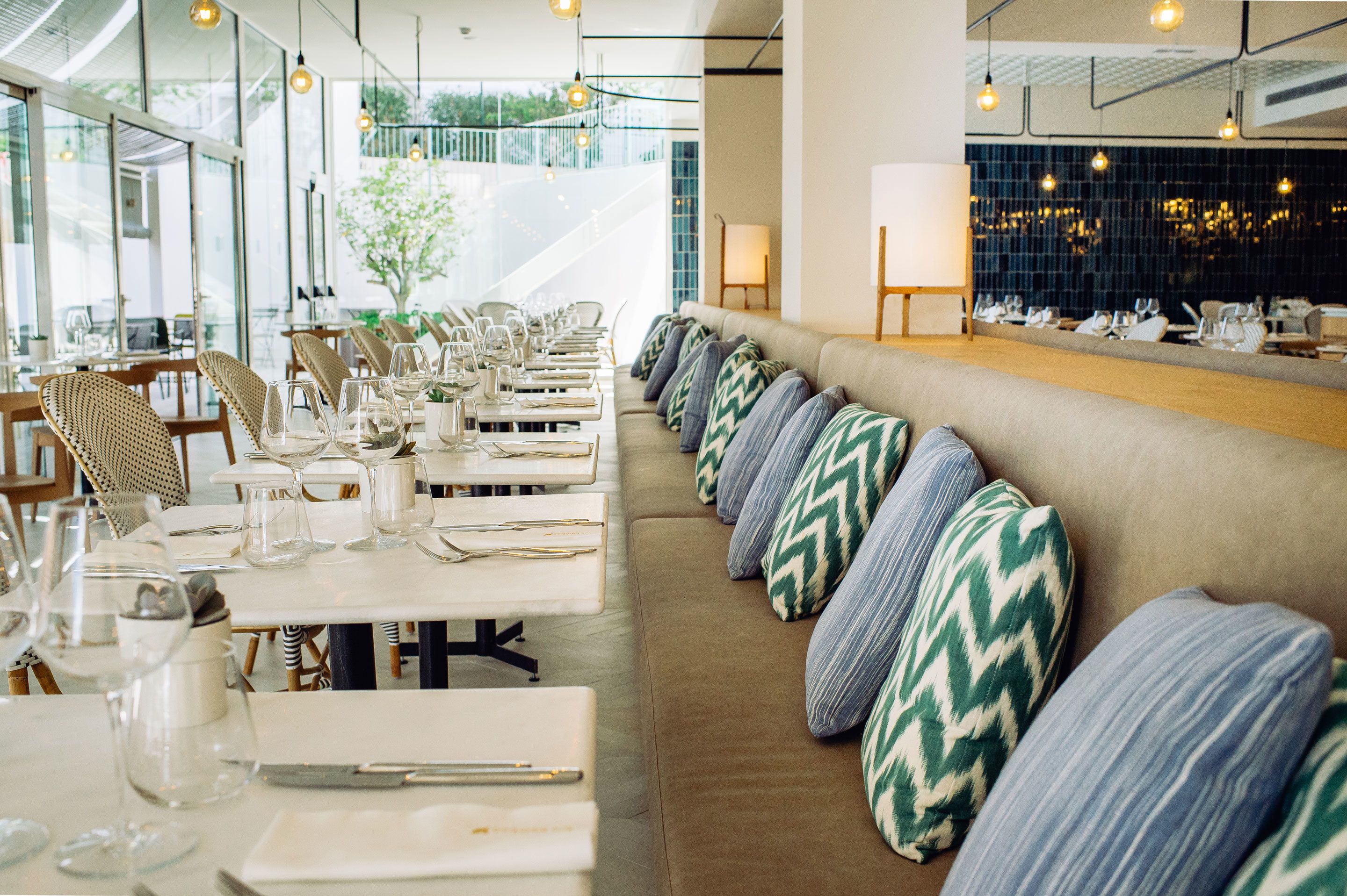 A restaurant with long communal tables, cushioned seating, and hanging pendant lights.