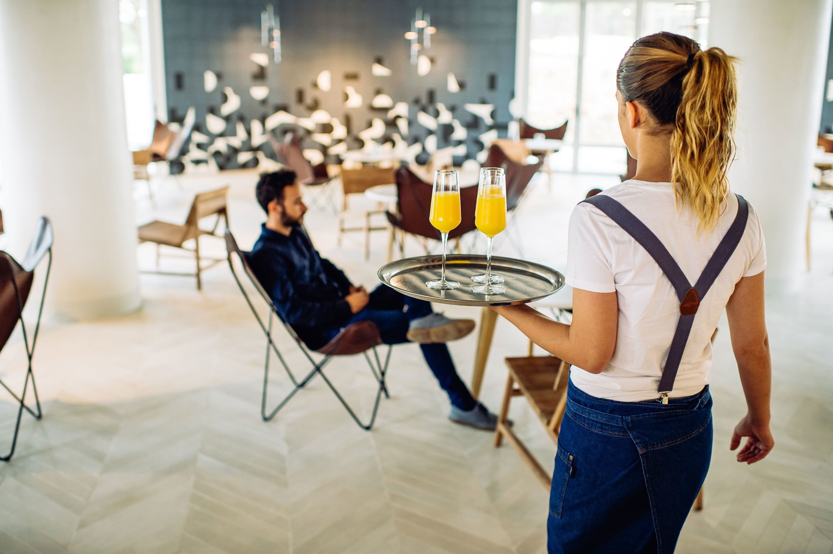 Une personne tenant un plateau avec deux verres de jus d’orange dans un espace intérieur moderne et bien éclairé, avec des chaises et des tables.