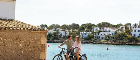 A coastal scene with a couple on bicycles, a stone building, and white houses in the background.