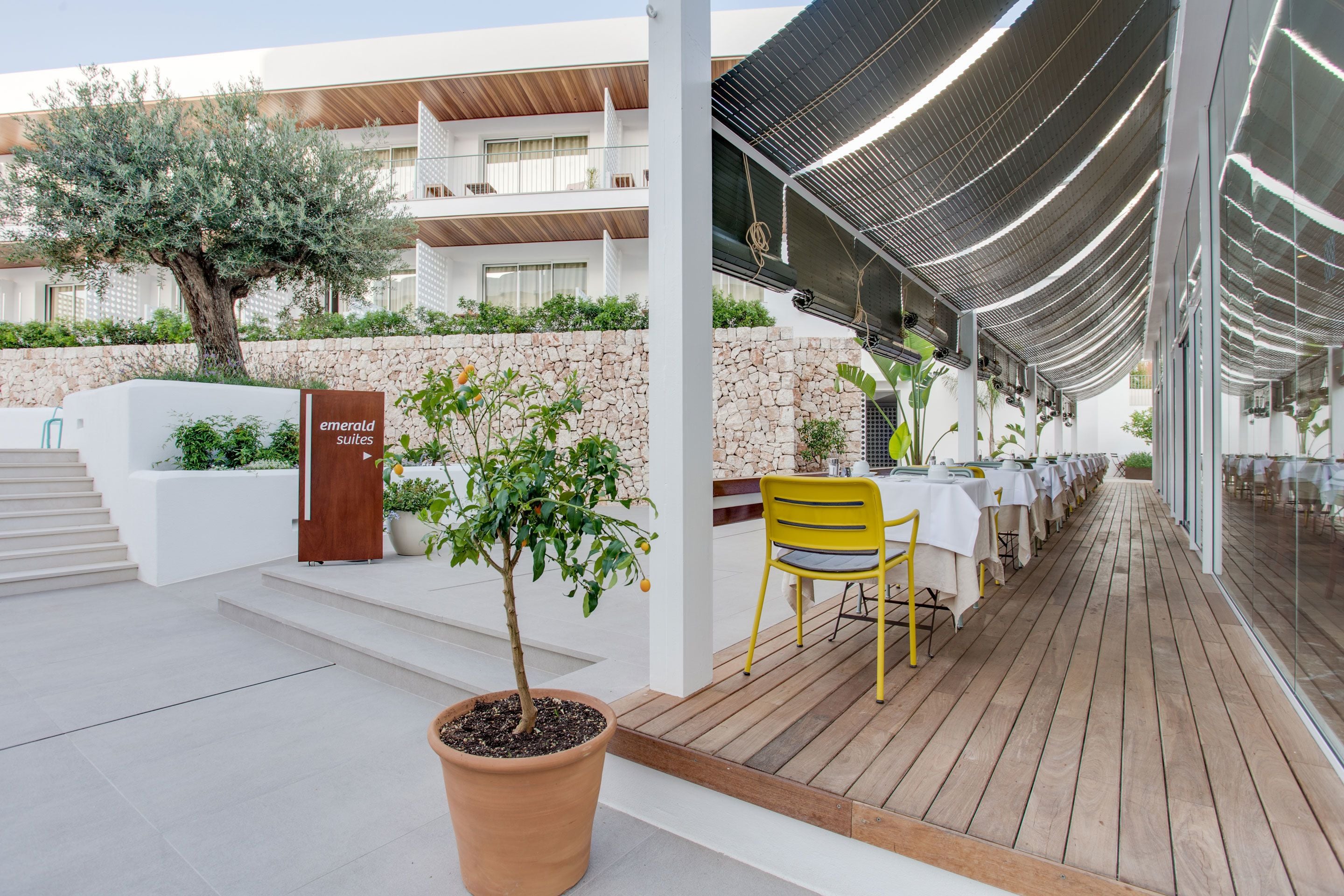Un espacio moderno para comer al aire libre con terraza de madera, sillas amarillas y mesas blancas. Se ve un árbol grande y una planta en maceta.