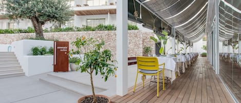 Un espacio moderno para comer al aire libre con terraza de madera, sillas amarillas y mesas blancas. Se ve un árbol grande y una planta en maceta.