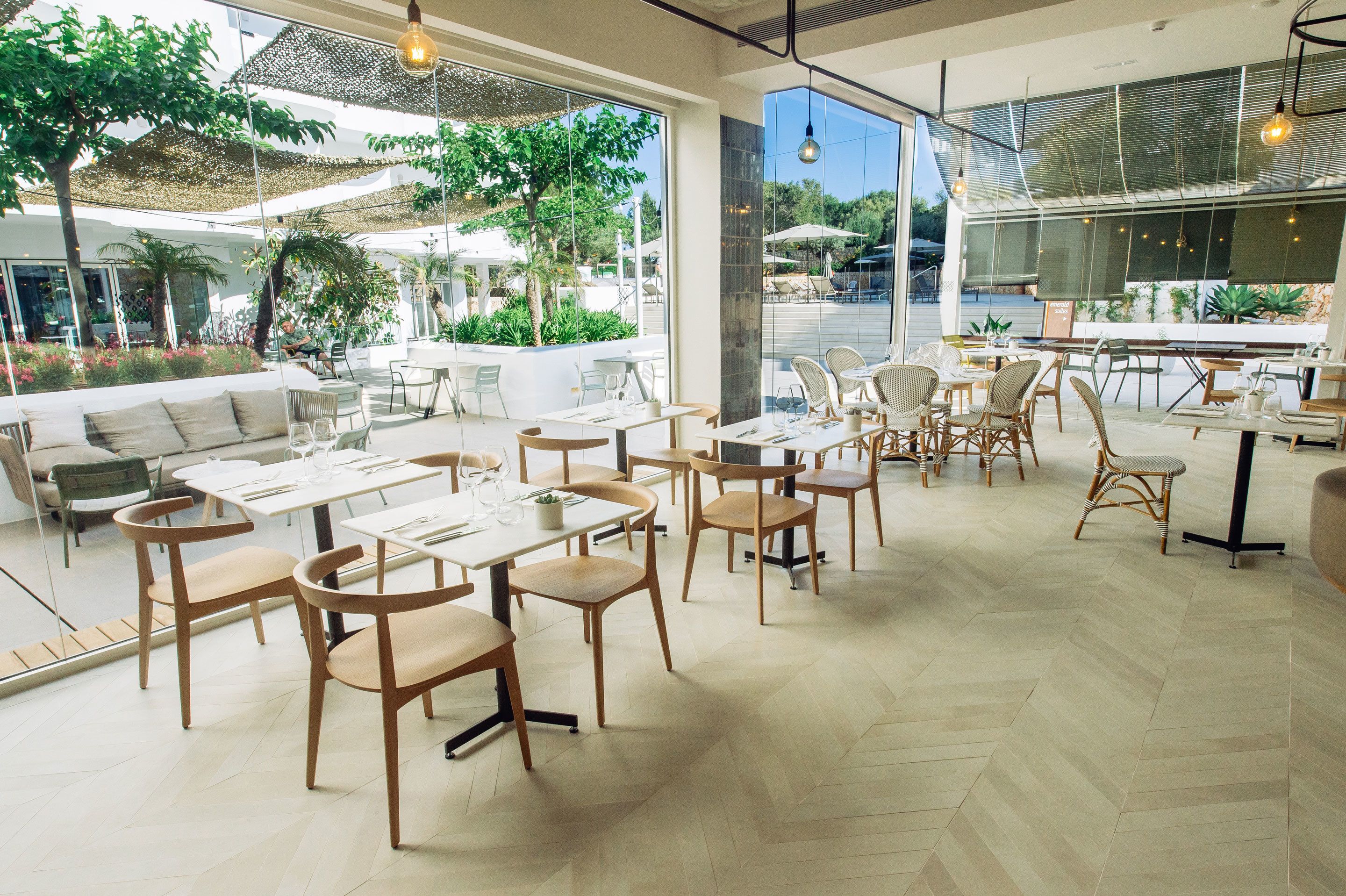 A spacious outdoor dining area with multiple tables and chairs, surrounded by large windows offering views of the outdoors.