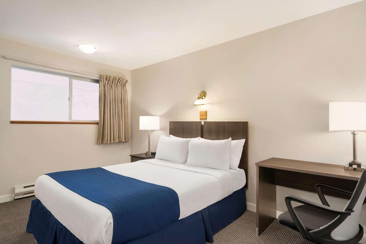 Ein Hotelzimmer mit einem großen Bett, einem Schreibtisch und einem Stuhl. Ein Fenster mit Vorhängen, eine Schreibtischlampe und eine Wanduhr sind ebenfalls vorhanden.