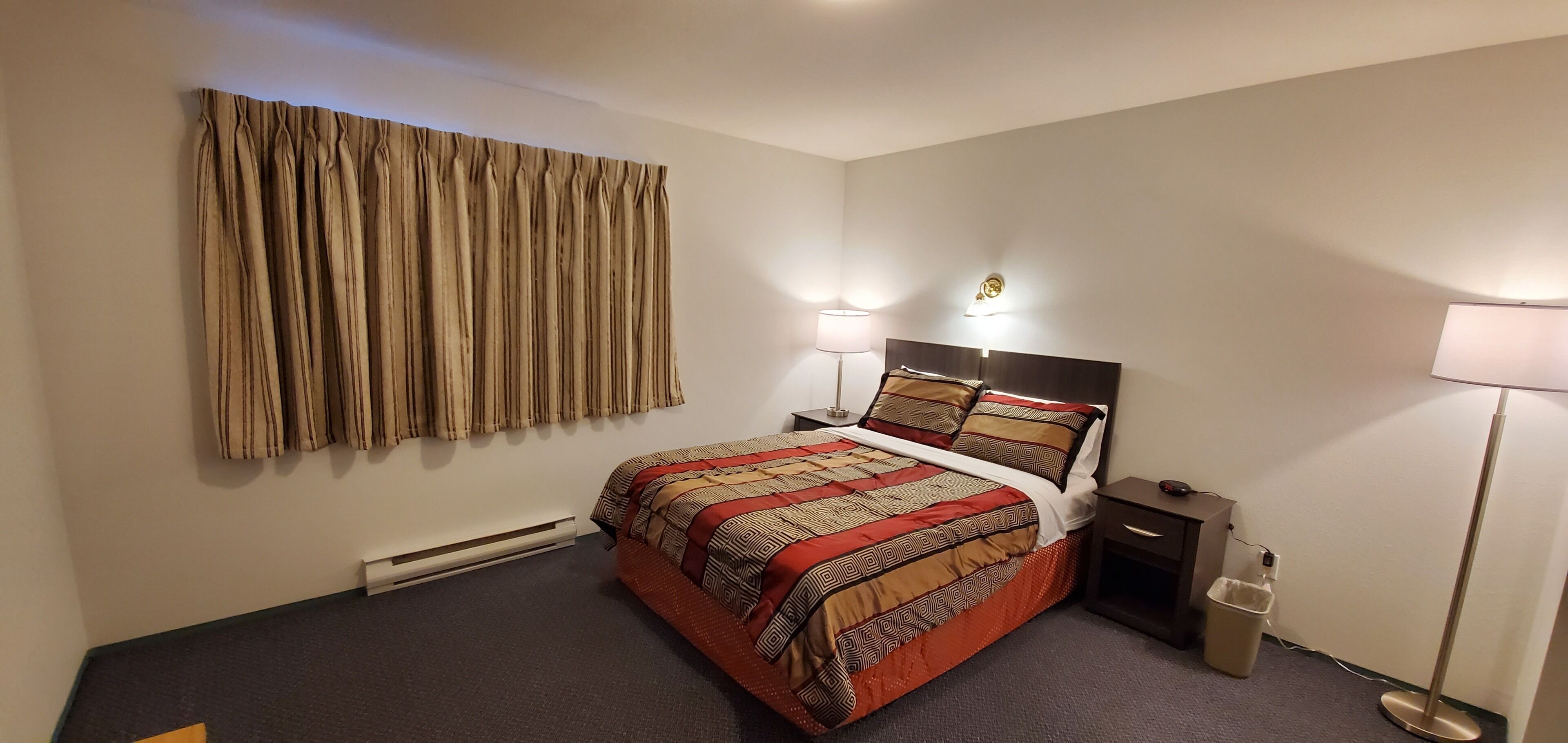 A hotel room with a bed, two bedside lamps, a nightstand, a floor lamp, a wall-mounted light, and a window with curtains.