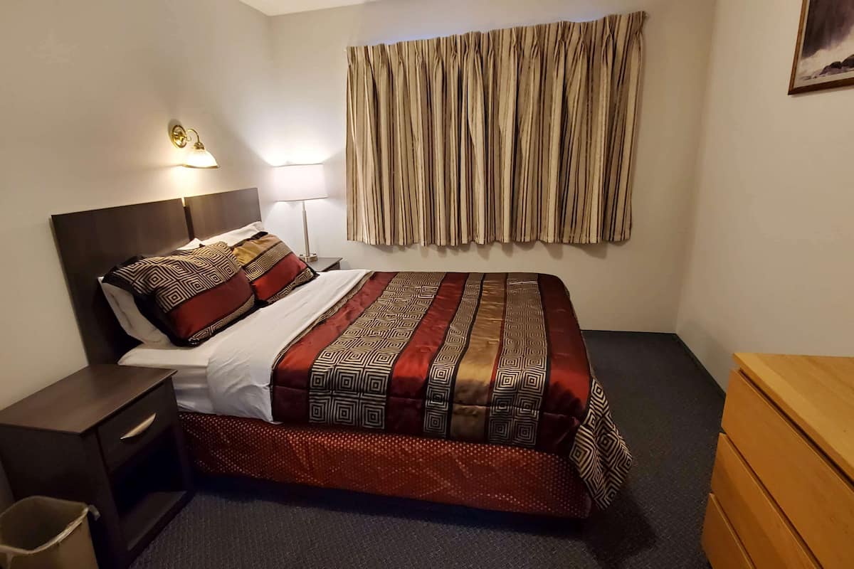 Ein Hotelzimmer mit Bett, Nachttisch, Lampe, Holzkommode und einem Fenster mit Vorhängen.