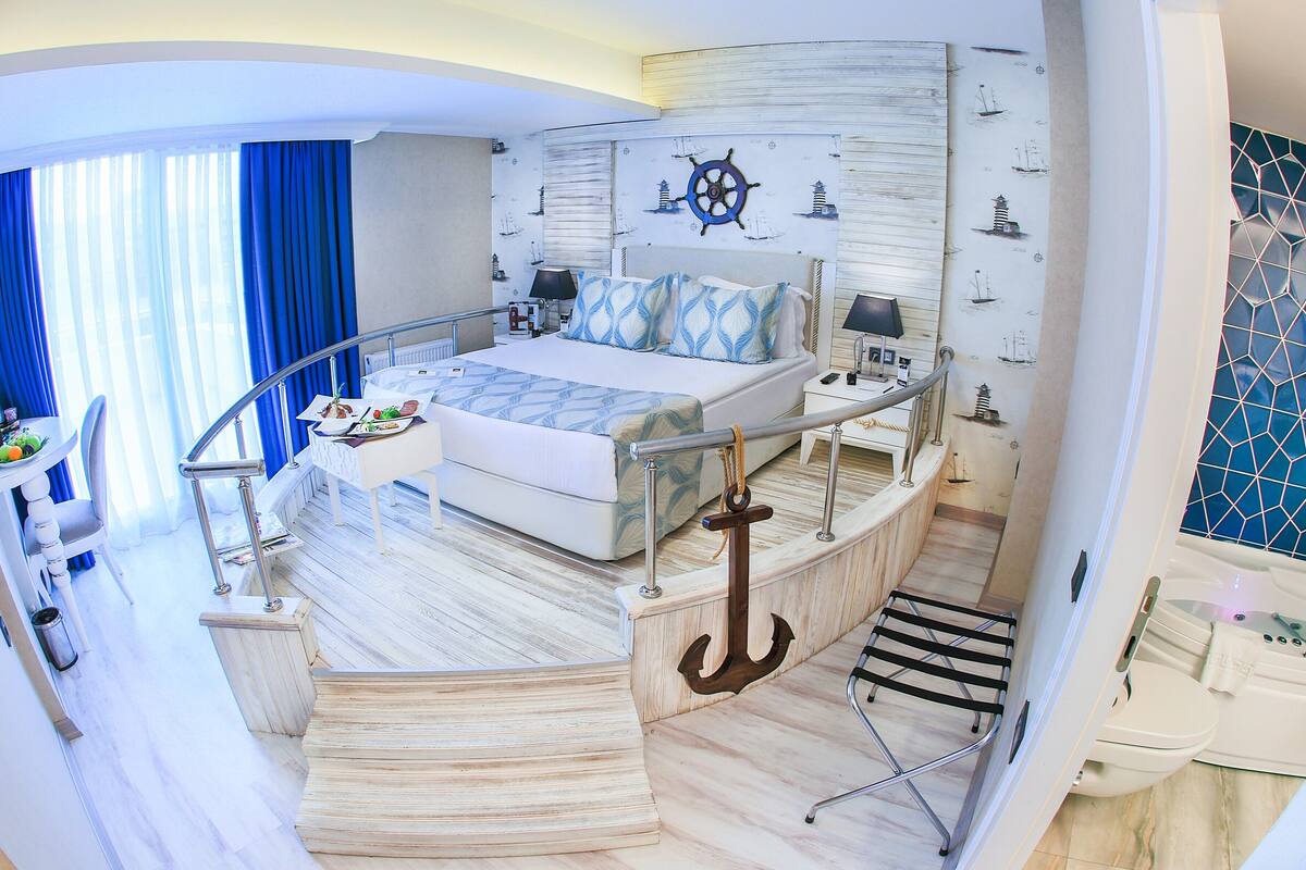 A nautical-themed hotel room with a bed, a small desk, a chair, a window with curtains, and a ship's wheel decoration.