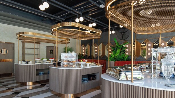 Un moderno hall de hotel con tres barras para desayuno, cada una con un diseño geométrico único y detalles dorados. El espacio presenta un patrón de piso a cuadros y una gran planta verde al fondo.