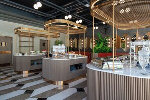 A modern hotel lobby with three round breakfast bars, each with a unique geometric design and gold accents. The space features a checkered floor pattern and a large green plant in the background. - Demonti Otel (Ankara)